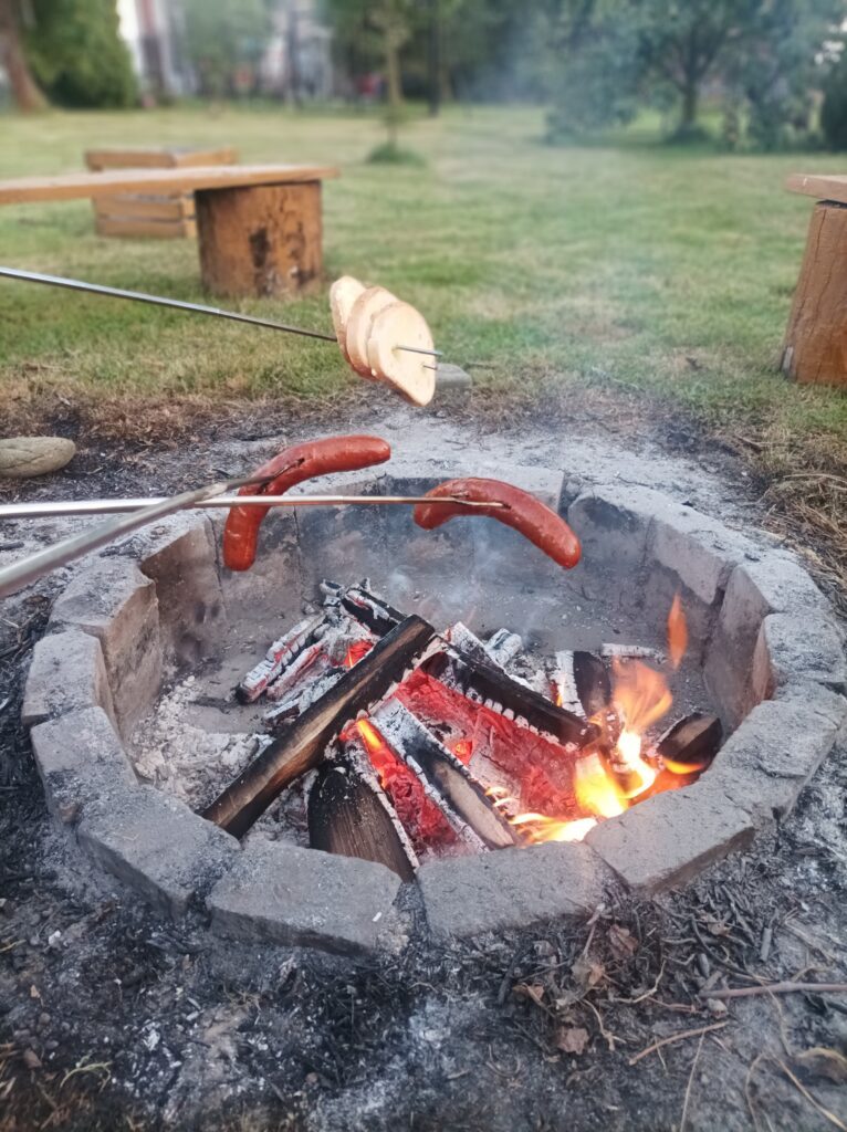 Palenisko w ogrodzie z trzema kiełbaskami pieczonymi na patykach, przygotowane w tradycyjny sposób nad ogniskiem