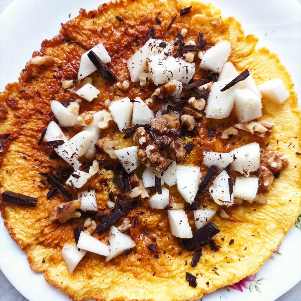 Na zdjęciu widnieje apetyczny omlet podany na talerzu. Jego złocista powierzchnia jest delikatnie posmarowana miodem, a na wierzchu ułożone są cienkie plasterki gruszki. Całość posypana jest kawałkami orzechów włoskich oraz startą gorzką czekoladą, która zaczyna się lekko topić, tworząc pyszny deserowy akcent. Omlet wygląda na ciepły i zachęcający, gotowy do podania.
