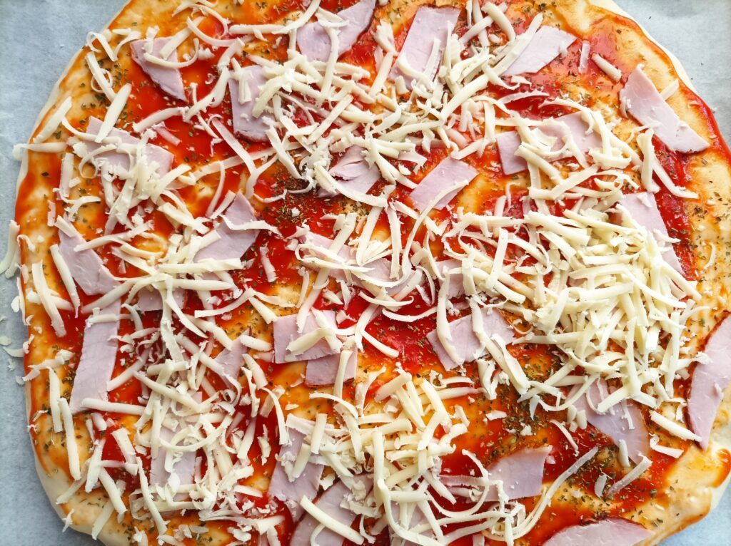 Pizza na grubym cieście posmarowana przecierem pomidorowym i posypana startym oregano ora ser mozzarella i szynka